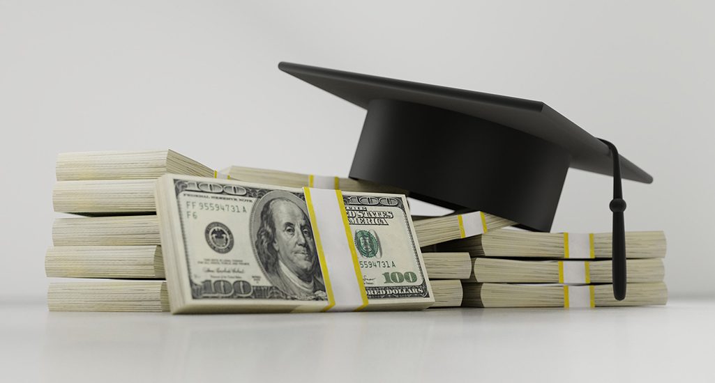 Several wads of hundred dollar bills with a graduation cap resting on top