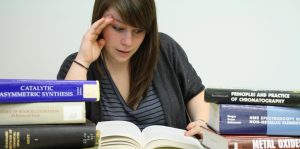 Student with stressed expression reading book, surrounded by stacks of other books