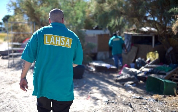 Two men wearing LAHSA shirts in a yard with items belonging to those experiencing homelessness
