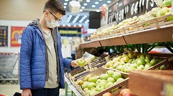 A masked shopper standing in the fruit section of the grocery store holds a green apple.