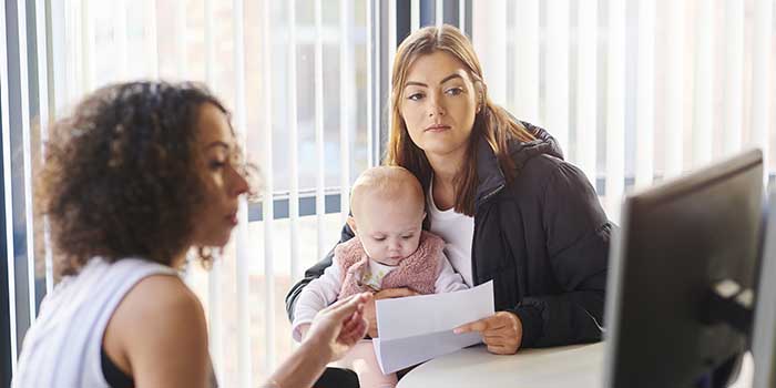 One woman is speaking to another woman who is holding an infant and a letter. The first woman is explaining something on the computer screen in front of them.