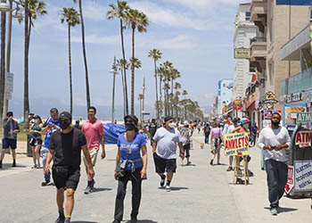 People walking on Venice Beach boardwalk during pandemic, most are wearing masks and there are palm trees in the background.