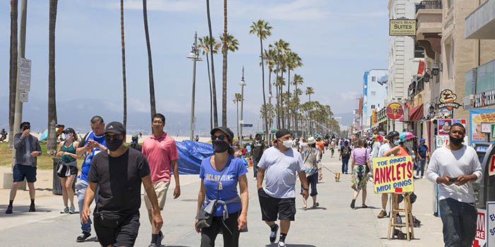People walking on Venice Beach boardwalk during pandemic, most are wearing masks and there are palm trees in the background.