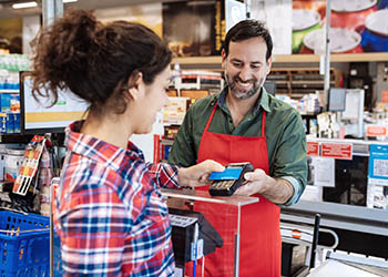 A younger woman holds her a payment card up to a cashier at a grocery store in the checkout line. The cashier is holding a card reader.