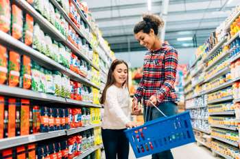 A mom and her young daughter are in a grocery store aisle. Both are smiling as the daughter places something into the hand basket the mom is holding