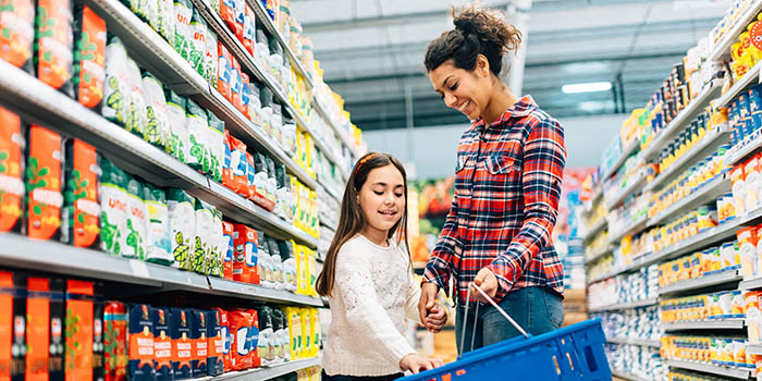 A mom and her young daughter are in a grocery store aisle. Both are smiling as the daughter places something into the hand basket the mom is holding