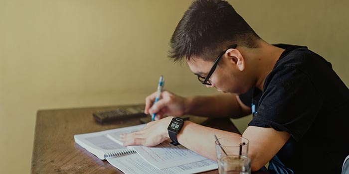A young adult male is sitting at a desk, writing in a notebook, with a calculator and iced coffee next to him.