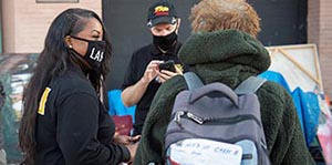 A person with a LAHSA face mask is speaking with a person who has a backpack on. Another person checks their phone in the background.