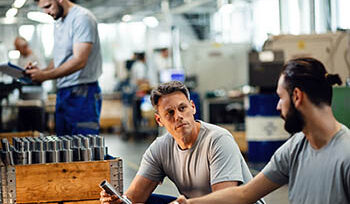 Two men, aged in their 30s and 40s, are kneeling in a factory, speaking with each other, and holding steel cylinders. Another man stands in the distance, checking a list on a clipboard.