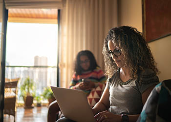 A woman in her mid 40s is sitting on a couch looking at a laptop. There is a girl in the background looking at something in her hands, and there is a balcony behind the girl.