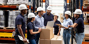A group of warehouse workers with hardhats on listens to a manager speaking with a laptop in front of him.