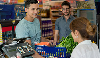 A younger man is smiling and checking out at a grocery store, you can see the cashier, a basket of food, and another person waiting in line.