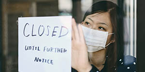 A woman with a face mask is placing a "CLOSED UNTIL FURTHER NOTICE" sign on a glass door, indicating an increase in unemployment.