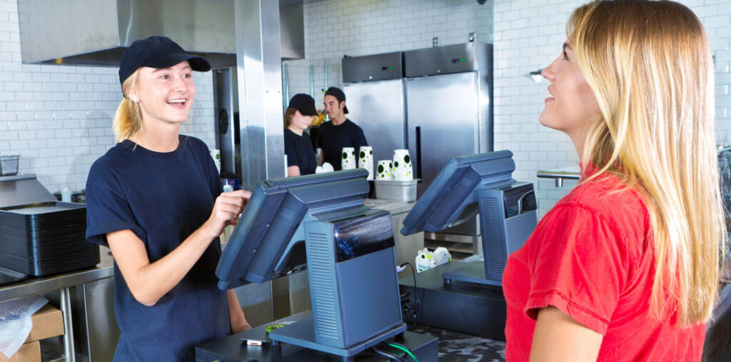 A young woman types in an order from a customer placing her order at a fast food restaurant. There are two more employees in the background.