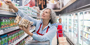 An older woman worker is standing on a ladder to put products on a shelf.