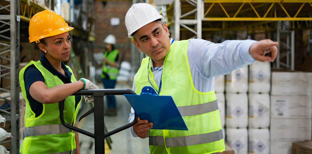 A younger woman listens to an older man who is holding a clipboard and pointing. They're in a warehouse, with hardhats on and green safety vests.
