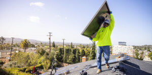 A solar installer hoists a panel as he walks along a roof with blue sky in the background.