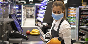 A younger woman cashier, wearing a face mask, scans an item at a grocery store.