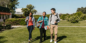 A group of 3 college students walk across a green lawn, smiling.