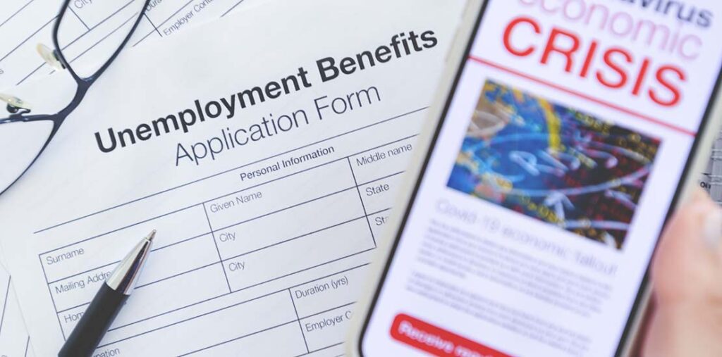 Image shows a unemployment benefits application form, glasses, a pen, and cell phone with news article that says "coronavirus economic crisis"