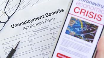 Image shows a unemployment benefits application form, glasses, a pen, and cell phone with news article that says "coronavirus economic crisis"