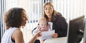 One woman is speaking to another woman who is holding an infant and a letter. The first woman is explaining something on the computer screen in front of them.